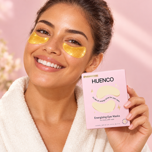 Smiling woman wearing gold under-eye gel patches and holding a pink HUENCO eye mask box.