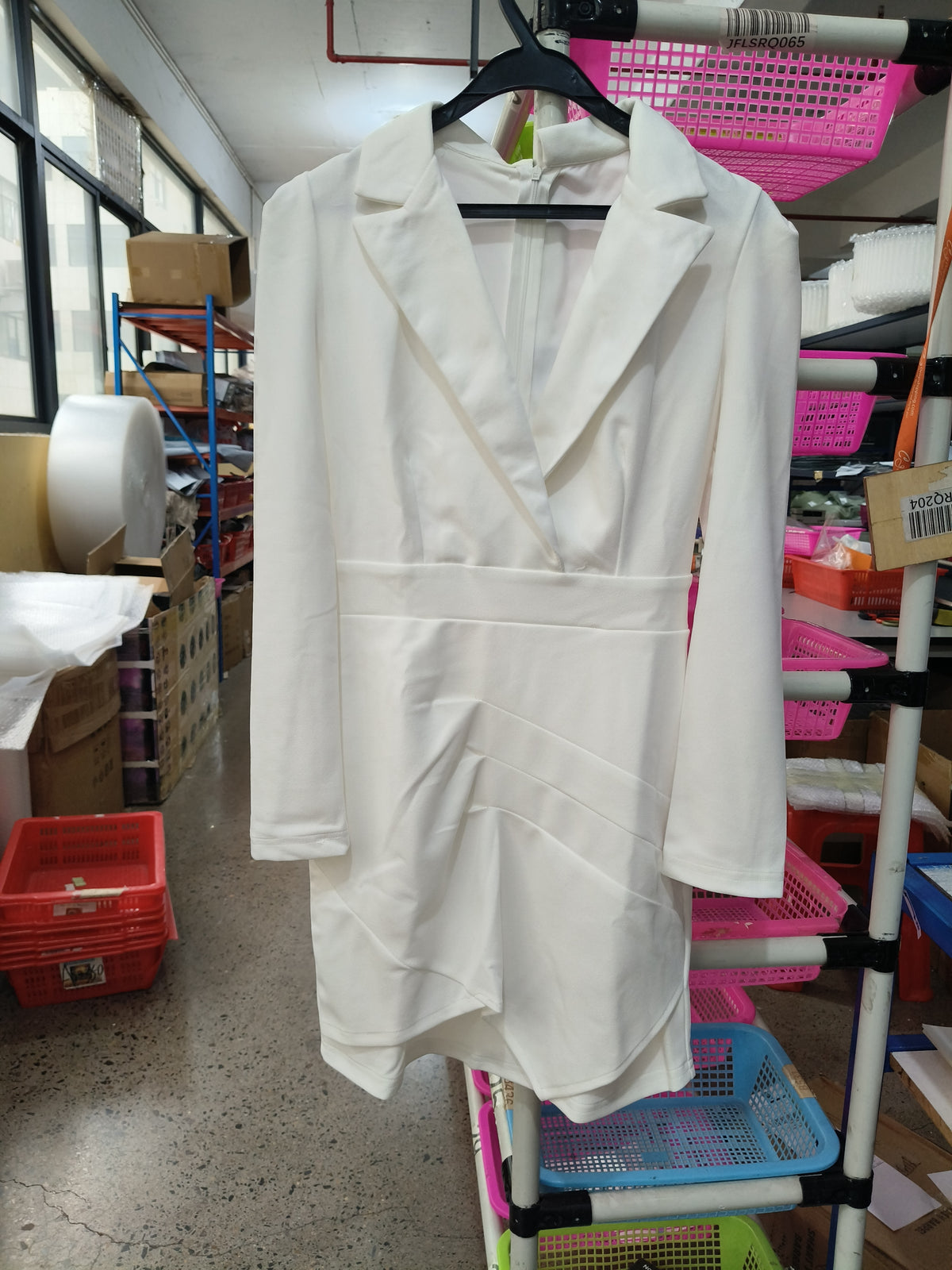 A white long-sleeve blazer-style dress with a structured collar and draped front detail, hanging on a black hanger in a storage or retail workspace with shelves and bins visible in the background.