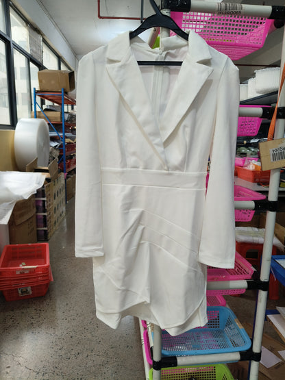 A white long-sleeve blazer-style dress with a structured collar and draped front detail, hanging on a black hanger in a storage or retail workspace with shelves and bins visible in the background.