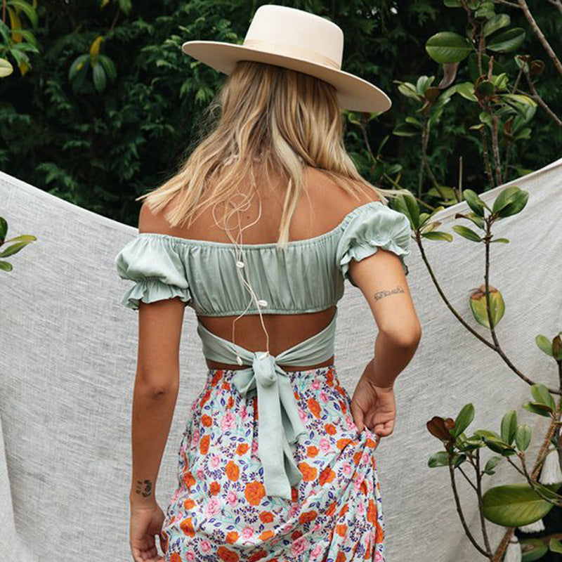 Woman wearing a light green off-shoulder top and floral skirt with a wide-brimmed hat, standing outdoors.
