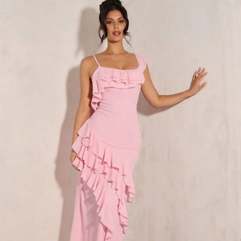 A woman modeling a light pink ruffled dress with spaghetti straps and an asymmetrical layered hem, posed against a textured off-white wall.
