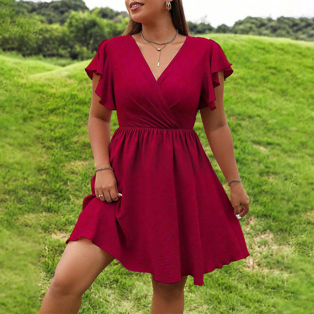 A woman wearing a short magenta wrap-style dress with flutter sleeves, standing outdoors on a grassy hill.