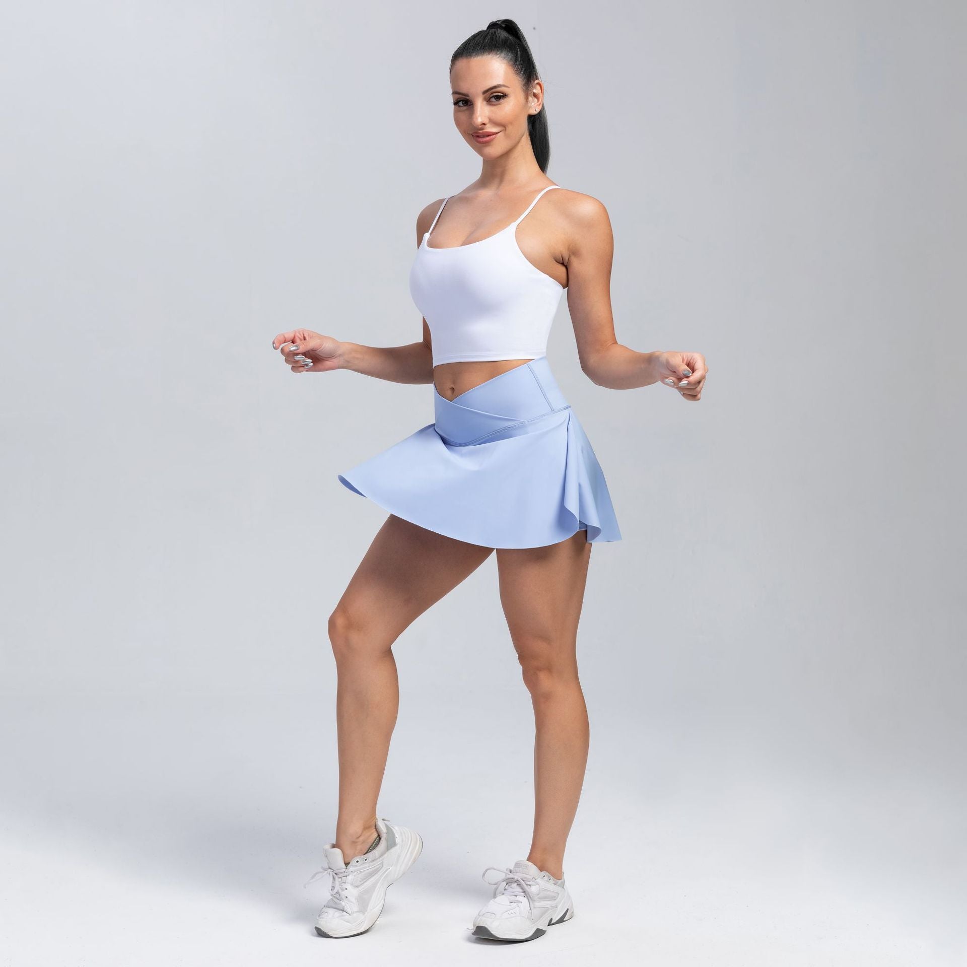 A woman modeling a white sports bra and a light blue athletic skirt, paired with white sneakers, against a light gray studio background.