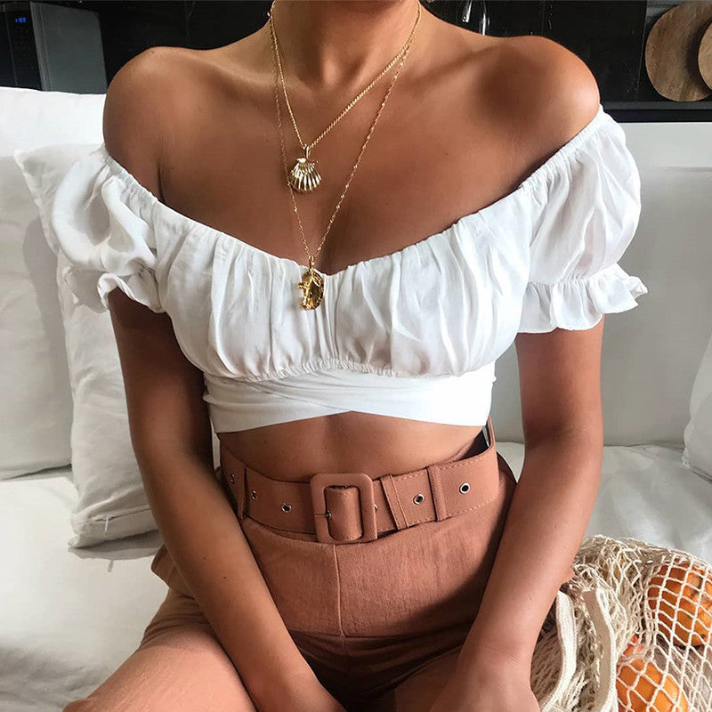 woman wearing a white off-shoulder top and brown pants sitting on a couch.