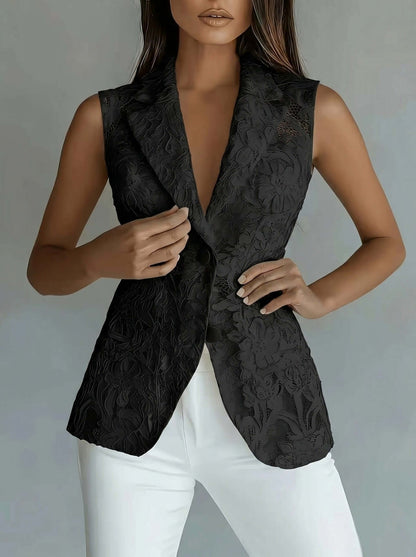 A woman wearing a sleeveless black textured blazer vest with a deep V-neck and white pants, shown from the neck down against a neutral gray studio background.